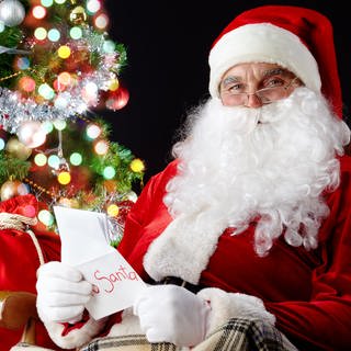 Der Weihnachtsmann vor einem Weihnachtsbaum mit einem Sack Geschenke und einer Weihnachtskarte
